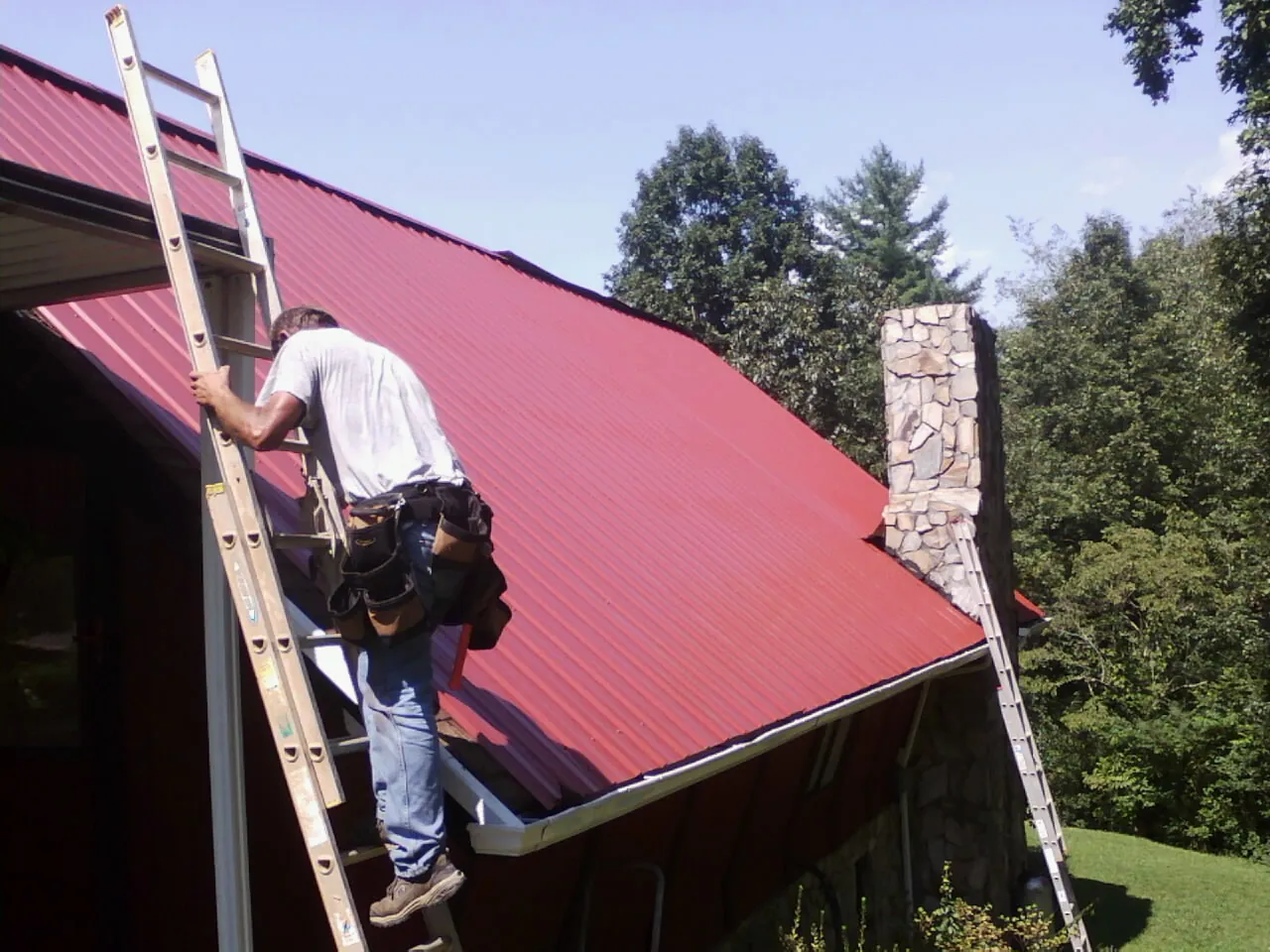 Roofer on a ladder beside a red metal roof and stone chimney — Soffit & Fascia Repair by Roofing Brothers in Terre Hill